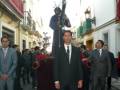 Una procesión religiosa con personas vestidas formalmente, incluyendo un hombre en traje negro con una armadura y un hombre en traje gris.