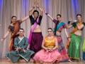 Una escena de una danza tradicional indígena, con bailarines vestidos en trajes coloridos y complejos. La imagen muestra a cinco bailarinas, tres de ellas en trajes tradicionales indios y dos vestidos con un estilo más moderno. La escena está iluminada por luces azules y el fondo es un telón de color beige.