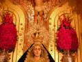Imagen de una Virgen María vestida con un manto negro y tocado dorado, rodeada por flores rojas en una iglesia.