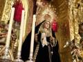 Imagen de una figura religiosa vestida de negro, posiblemente una virgen, con un altar dorado y una cruz en el fondo.