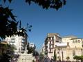 Una plaza con una fuente central y edificios históricos en el fondo, rodeada de árboles y flores.