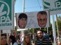 Un hombre sostiene un cartel con una caricatura de Maradona y otro de Rajoy, junto a imágenes de euros. El cartel dice "NOS ROBAN". Detrás, se ven otros carteles y personas con pancartas.