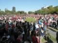 Una multitud de personas se reúnen en un parque, algunos con pancartas y banderas rojas. La escena está iluminada por el sol, con árboles y edificios al fondo.