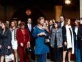 A una mujer anciana con vestido azul y bufanda blanca, seguida por varias personas en trajes formales. La escena parece ser una ceremonia o evento oficial, posiblemente en España, dada la presencia de vestimenta tradicional y el estilo arquitectónico.