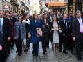 Una multitud camina por una calle con edificios de fondo. La imagen muestra a varias personas vestidas formalmente, algunas de ellas con chaquetas y otros con trajes. La gente parece estar en un evento o reunión importante, dado el ambiente formal y la presencia de personas con trajes.