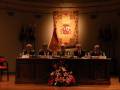 Una reunión formal en un salón con una bandera española y un escudo de armas en el fondo. Los asistentes se sientan alrededor de una mesa con flores y documentos en frente.