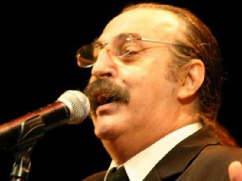 Un hombre con bigote y gafas, vestido de traje negro, canta en un escenario con un micrófono.