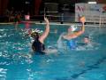 Un equipo de waterpolo en acción, con un jugador lanzando la pelota hacia el arco mientras otros jugadores se preparan para reaccionar. La piscina está iluminada con luces rojas y amarillas, y hay un banner de Andalucía en el fondo.