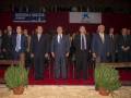 Un grupo de hombres en trajes formales se encuentran frente a un escenario con carteles promocionales. La imagen parece ser de una ceremonia o evento formal, posiblemente un congreso o reunión empresarial.