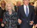 Una mujer mayor con cabello blanco y una abrigo de piel camina junto a un hombre en traje, ambos sonríen y parecen estar en una celebración.