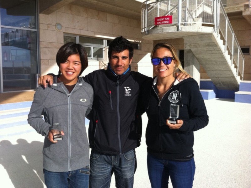 Tres personas sonrientes posan con trofeos en una escena al aire libre, posiblemente en un evento deportivo.