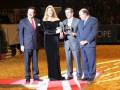 Una celebridad en un vestido negro y dos hombres en trajes azules están recibiendo un premio. La celebridad sostiene una estatuilla mientras los hombres la miran con atención. El fondo muestra un escenario con una bandera roja y blanca, probablemente en un evento formal o de premiación.