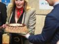 Una mujer con cabello largo y rubio, usando un abrigo beige y una bufanda roja, está recibiendo un pastel de chocolate con crema. Un hombre le ofrece el pastel con una mano. Detrás de ellos, hay carteles publicitarios y un edificio de ladrillo.