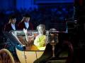 Una mujer en un elegante vestido amarillo se sienta en el asiento delantera de una carroza, mientras dos hombres vestidos formalmente la acompañan. La escena parece ser parte de una ceremonia o evento importante, con un público en el fondo.