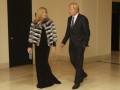 Una mujer vestida con un abrigo de piel y una falda larga negra camina junto a un hombre en traje negro. Ambos parecen estar en una sala de recepción o evento formal, con un fondo oscuro y luces brillantes.