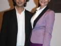 Dos personas posan juntas en un evento formal. La persona de la izquierda lleva una chaqueta negra y pantalones vaqueros, mientras que la persona de la derecha está vestida con un traje de color púrpura y una camisa blanca.