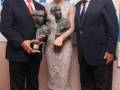Tres personas posan con trofeos en un evento formal. La imagen muestra a una mujer en el centro, rodeada por dos hombres mayores que sostienen trofeos. El fondo muestra un cartel con texto en idioma español, probablemente el nombre de una organización o evento.