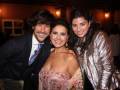 Tres personas sonrientes en un evento formal, posando juntas. El hombre a la izquierda lleva una chaqueta con rayas y un corbata, mientras que las dos mujeres a la derecha llevan vestidos elegantes. El fondo muestra una decoración de lujo con velas y un ambiente festivo.