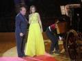 Una mujer en un vestido amarillo y un hombre en traje azul se encuentran junto a una carroza de caballo. Un hombre con una camiseta roja y pantalones negros está sentado en la carroza. El escenario parece ser una alfombra roja con un fondo de madera y lona.