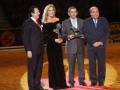 Una mujer en un vestido negro brillante sostiene un trofeo mientras está rodeada por tres hombres en trajes formales. La escena parece ser un evento de premiación o reconocimiento, con una alfombra roja y un escenario iluminado en el fondo.