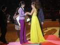 Una mujer en un vestido amarillo entrega un premio a una mujer en un vestido púrpura, ambos están sobre un escenario rojo y amarillo.