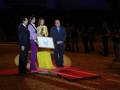 Una escena formal con cuatro personas en un escenario iluminado, dos hombres y dos mujeres. La mujer más joven sostiene una placa de reconocimiento mientras los otros tres miran hacia ella. La escena parece ser parte de un evento formal, posiblemente un premio o reconocimiento.