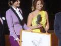 Premio entregado en una ceremonia formal, ambas mujeres sonríen mientras una sostiene un reconocimiento y la otra un premio.