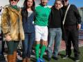 Una persona con una chaqueta de piel, otra con una blusa azul y botas marrones, un futbolista en camiseta verde y pantalón blanco, y dos hombres con chaquetas negras. Todos están en un campo de fútbol, posando para la foto.