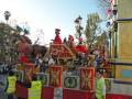 Una carreta festiva con un elefante en el centro, rodeado de personas vestidas como personajes navideños. La carreta está decorada con banderas y un árbol de Navidad en el fondo.