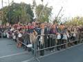 Una multitud de personas se reúne en una calle, detrás de un corredor de metal. La gente está vestida con abrigos y sombreros, algunas llevan bolsas de compras. El ambiente es festivo y el cielo está claro.