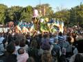 Un carnaval en pleno apogeo, con disfraces coloridos y figuras gigantes en el centro. La multitud de espectadores observa atentamente la celebración, mientras los participantes se preparan para su actuación.