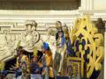 Imagen de una carroza real con personajes vestidos en trajes históricos, posiblemente de la época del reinado de un monarca. La carroza está decorada con símbolos y figuras antiguas, lo que sugiere un evento ceremonial o festivo.