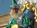 Un personaje vestido con trajes de fantasía, incluyendo un casco verde y una capa plateada, posando frente a un edificio con detalles dorados.