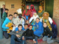 Un grupo de niños y adultos posan juntos en un ambiente festivo, con adornos navideños al fondo. Algunos niños llevan disfraces de los Reyes Magos, mientras otros vestidos con trajes tradicionales. La escena transmite alegría y celebración navideña.