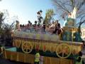 Un desfile de carros de flores en una ciudad, con personas vestidas de blanco y carros decorados con flores y figuras.