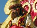 Un hombre vestido con trajes de gala dorados y rojos, sosteniendo un gran cetro dorado. El fondo es una estructura decorativa de oro y rojo con letras en relieve.