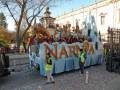 Un carnaval en Narnia, con un carro grande y colorido lleno de personajes y figuras, rodeado por niños vestidos con disfraces. La imagen muestra un ambiente festivo y lleno de color, con confeti cañoncillo en el suelo.