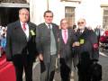 Tres hombres vestidos formalmente caminan por una calle con edificios blancos y puertas de metal. El primero lleva un abrigo negro con una corbata roja, el segundo tiene un abrigo verde con botones y una corbata azul, mientras que el tercero lleva un abrigo negro con una corbata roja y un bolso de mano. Todos llevan insignias amarillas en sus chalecos.