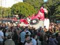 Una multitud disfruta de un desfile con una carroza en forma de dragón rosa y blanco, rodeada por árboles verdes.