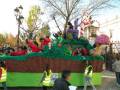 Un carnaval con un carro de gran tamaño decorado como una cesta de frutas. Los participantes están vestidos con chalecos amarillos y chaquetas rojas, mientras que el carro está lleno de frutas como manzanas y naranjas. La escena se desarrolla en un entorno urbano con árboles y una estructura de metal al fondo.