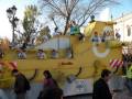 Desfile de carrozas en Sevilla, España, con un vehículo amarillo y detalles de la ciudad en el fondo.