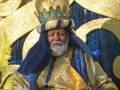 Un rey vestido con un traje dorado y azul, con un sombrero de copa y una corona en la cabeza. La imagen está iluminada con luces doradas y azules, creando un ambiente festivo.