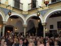 Una multitud asiste a una ceremonia en un edificio histórico con arcos y columnas de estilo neoclásico. La audiencia está sentada en filas, mirando hacia adelante con expresiones serias. La decoración interior incluye detalles dorados y luces colgantes, creando un ambiente formal y elegante.