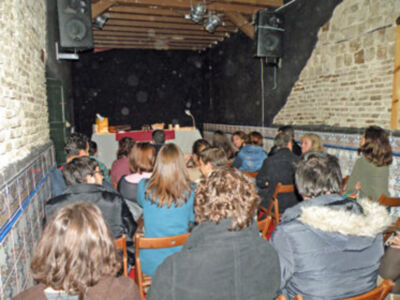 Una audiencia en un concierto, sentados en sillas de madera frente a una pared de ladrillos. La imagen muestra un ambiente acústico con luces y un micrófono en el escenario.