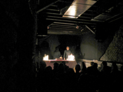Una persona en un escenario iluminado por lámparas, con una audiencia de siluetas en la oscuridad.