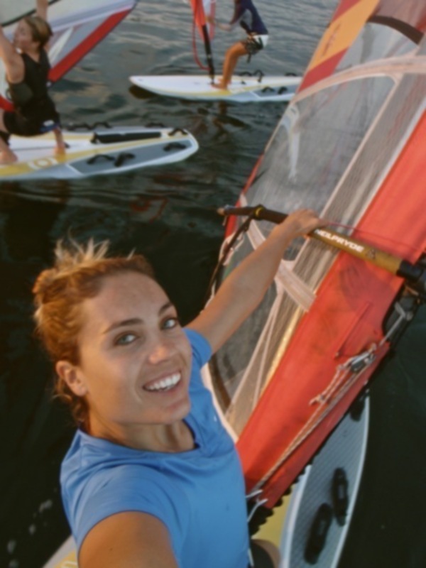 Una mujer sonríe mientras sostiene un paddleboard rojo y blanco en el agua. Detrás de ella, otros personas también están disfrutando del deporte acuático en el mar.