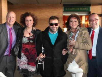 Una familia feliz posa para la foto en un ambiente familiar. La imagen muestra a cuatro personas, dos hombres y dos mujeres, vestidos formalmente. El hombre de la izquierda lleva un traje negro con una corbata roja, mientras que el hombre de la derecha lleva un traje azul con una corbata roja. La mujer de la izquierda lleva un vestido blanco con estampado floral y una corbata roja, mientras que la mujer de la derecha lleva una chaqueta beige con una bufanda colorida. Todos parecen estar en un...