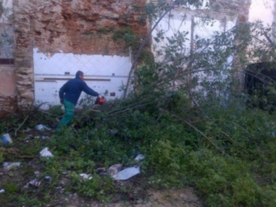 Un hombre con una chaqueta azul y pantalones verdes está cortando ramas caídas en un área con escombros y vegetación. La pared de atrás es roja y blanca, con un edificio adyacente visible a la derecha.
