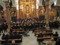 Una multitud de músicos militares toca en un templo con altares dorados y columnas, mientras que el público observa desde bancos.