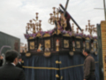 Imagen muestra una procesión con un carro de madera adornado con flores y cruces, acompañado por personas en trajes formales.